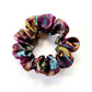 A liberty silk scrunchie with a multicolor paisley and floral pattern in purple, teal, yellow, and brown, shown on a plain white background.