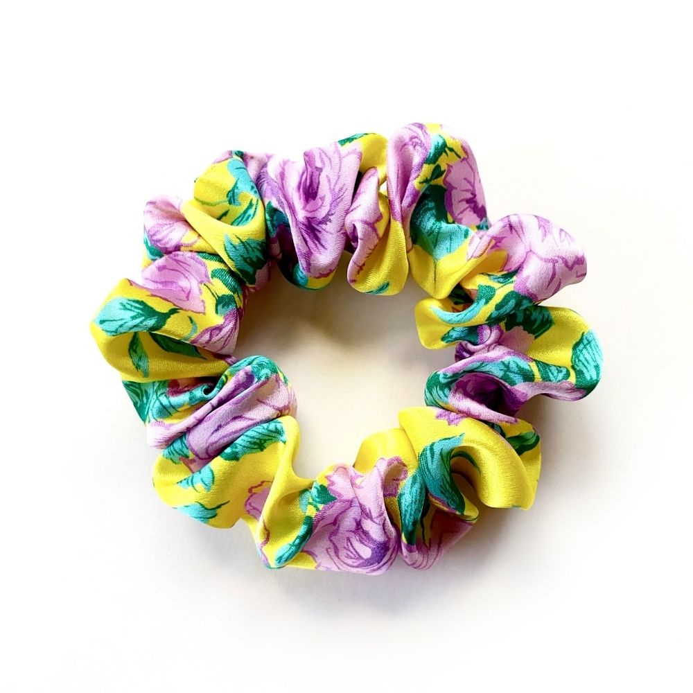 A liberty silk scrunchie with a vibrant floral print in yellow, purple, and green, photographed on a clean white background.