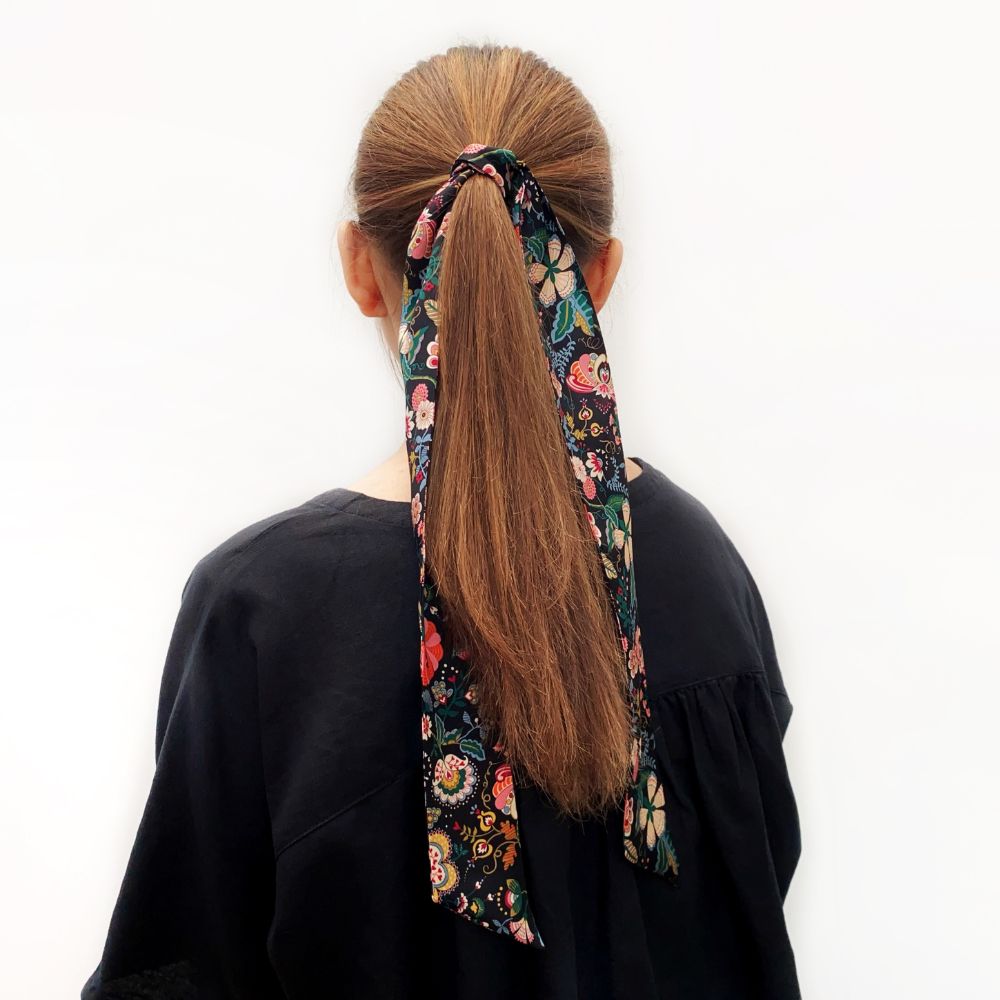 Person with a black Liberty floral print silk skinny hair scarf  tied in their ponytail on a white background