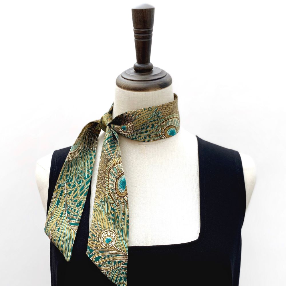 Mannequin wearing a black dress with a green and gold peacock feather patterned liberty silk skinny scarf around its neck on a white background