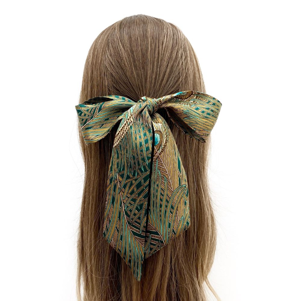 Liberty silk skinny scarf with green and gold peacock pattern tied into a hair bow  on a pony tail on white background