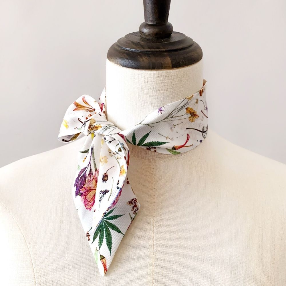 A white Liberty London silk skinny scarf with a colorful botanical and cannabis leaf pattern, tied in a knot and displayed on mannequin neck.