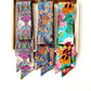 Three colorful patterned liberty london silk twill skinny scarves displayed side by side inside a light wood tray against a white background. The left tie has a pink, purple, and teal abstract pattern with a bird motif.