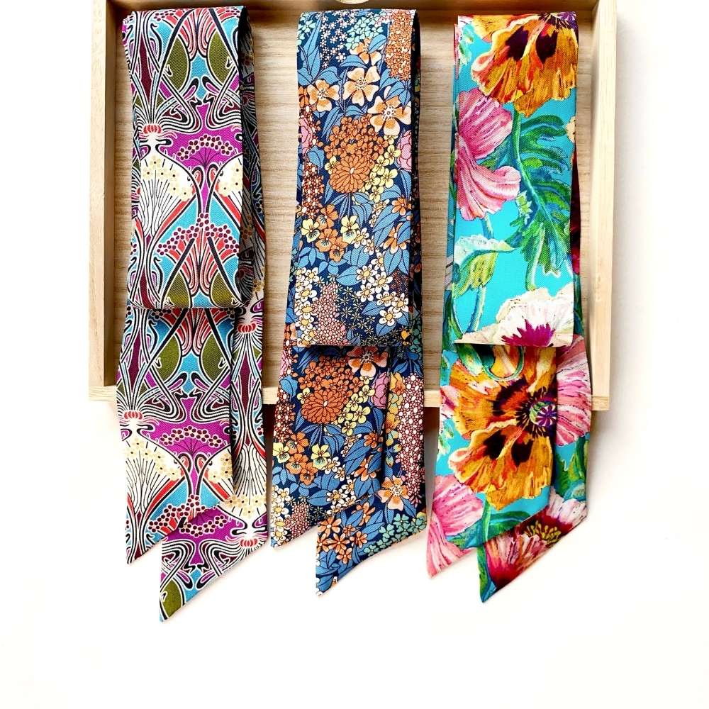 Three colorful patterned liberty london silk twill skinny scarves displayed side by side inside a light wood tray against a white background. The left tie has a pink, purple, and teal abstract pattern with a bird motif.