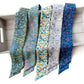 Five patterned Liberty London silk twilly scarves  in shades of blue, green, purple, and yellow are draped side-by-side on a light gray wooden tray against a white background