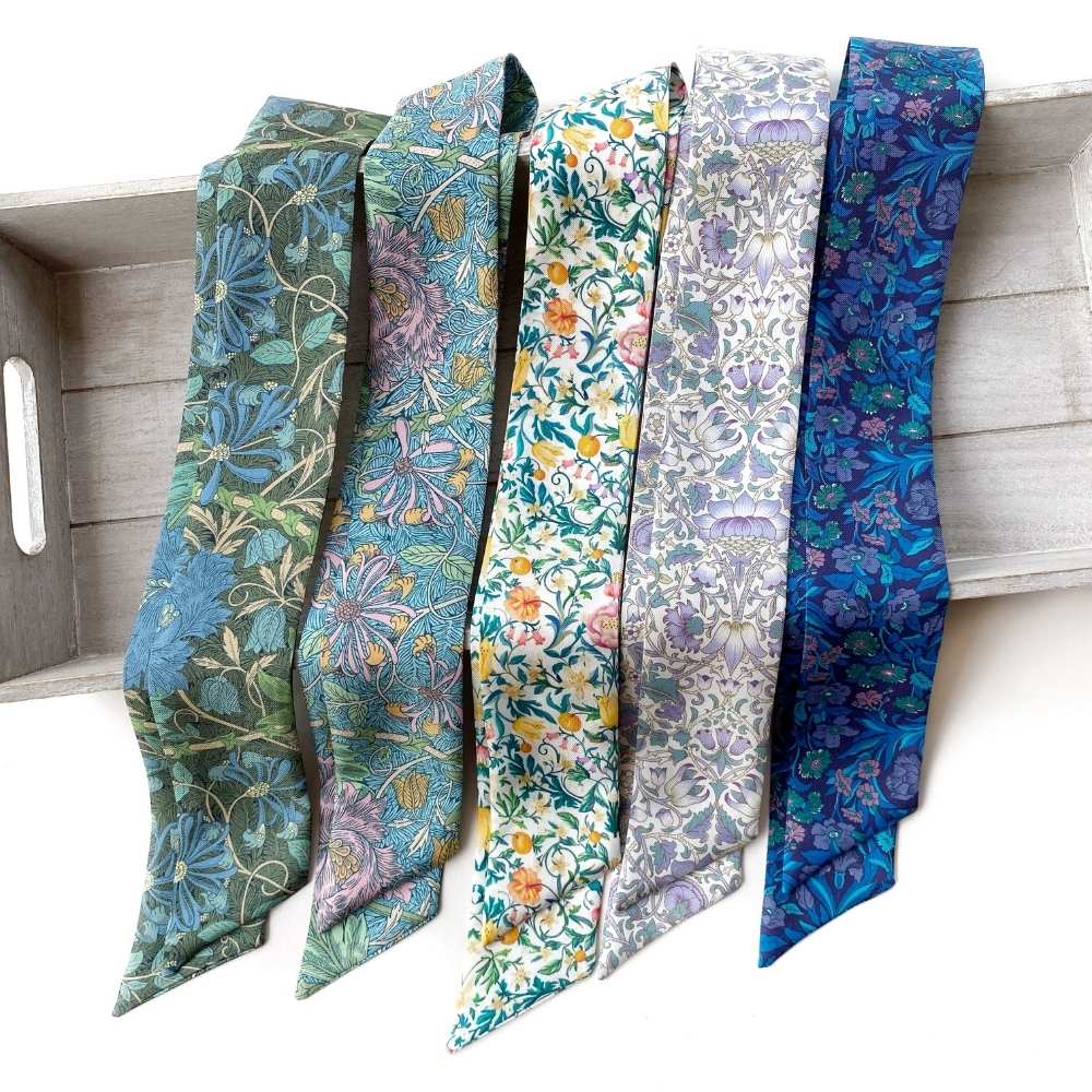 Five patterned Liberty London silk twilly scarves  in shades of blue, green, purple, and yellow are draped side-by-side on a light gray wooden tray against a white background