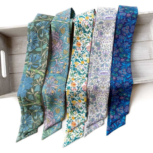 Five patterned Liberty London silk twilly scarves  in shades of blue, green, purple, and yellow are draped side-by-side on a light gray wooden tray against a white background