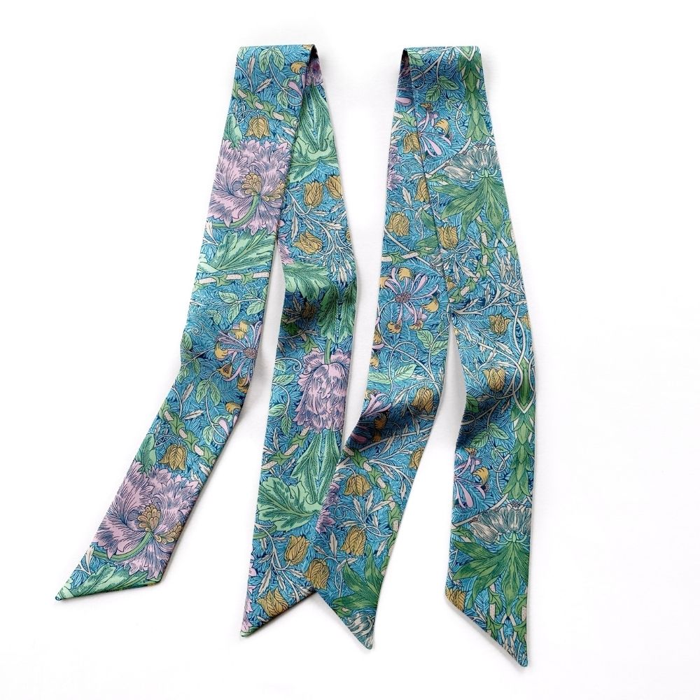 A long Liberty London silk twilly scarf with a dense botanical pattern featuring teal, green, purple, and gold tones, laid flat on a white background.