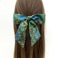 A Liberty London silk hair scarf with a blue and green floral pattern, tied into a bow on a person's brown hair against a plain white background.