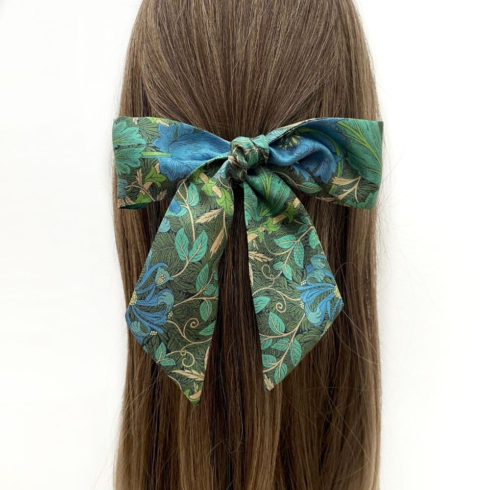 A Liberty London silk hair scarf with a blue and green floral pattern, tied into a bow on a person's brown hair against a plain white background.