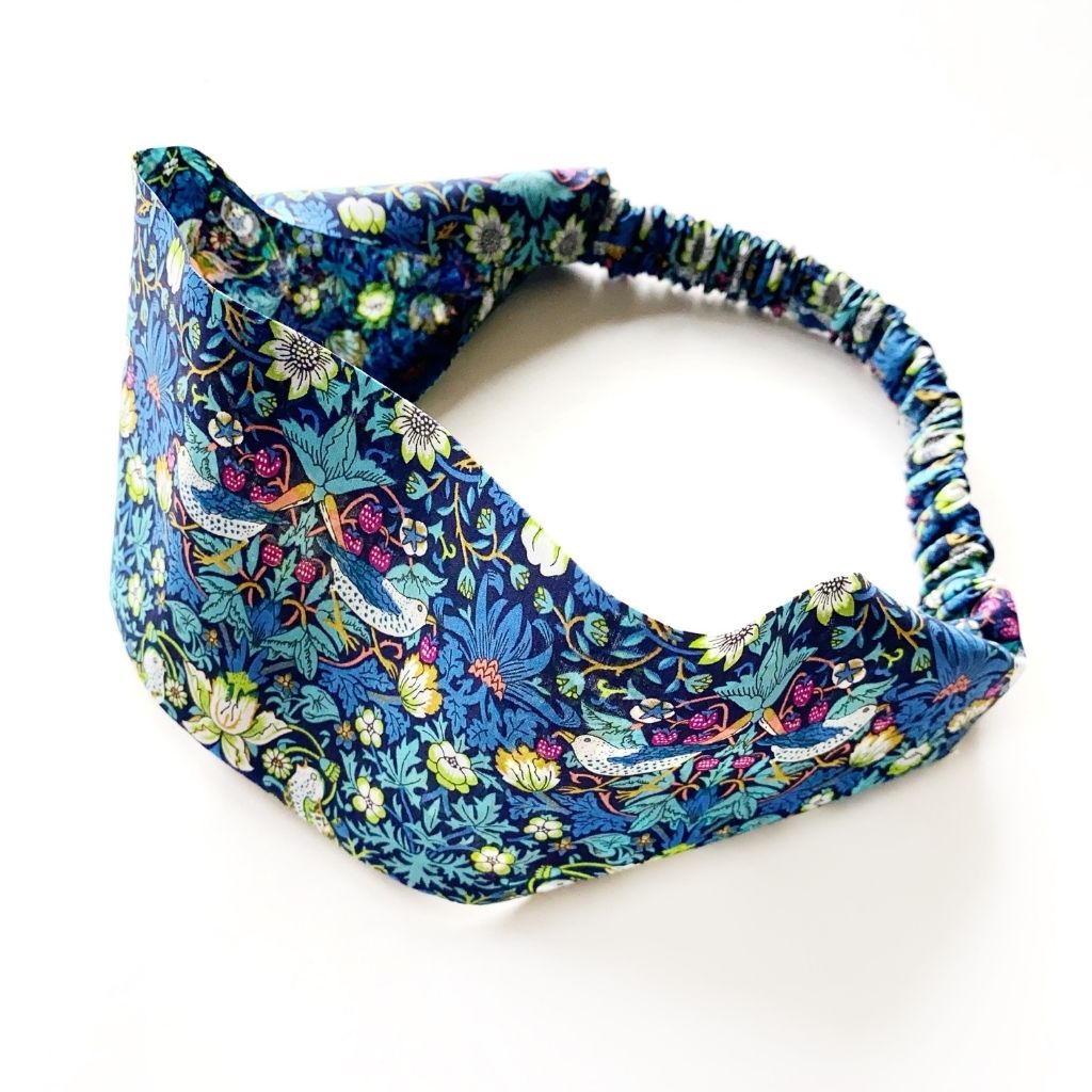 A Liberty London Tana Lawn cotton headband with an elastic back, featuring william morris print " strawberry thief" a detailed floral and bird pattern in blue, green, pink, and yellow on a white background.