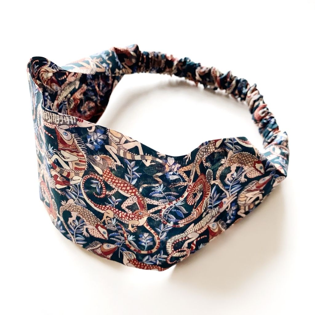 A Liberty London Tana Lawn cotton  headband with a gathered elastic back, featuring an intricate all-over animal print and floral motifs in navy blue, rust red, beige, and white, set against a plain white background.