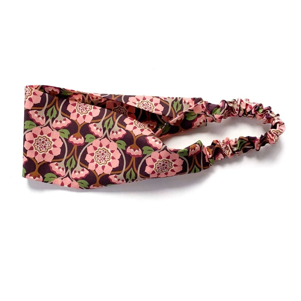 A Liberty London Tana Lawn cotton  headband with a pink and green floral pattern on a dark brown background, featuring an elastic band, shown on a plain white background.