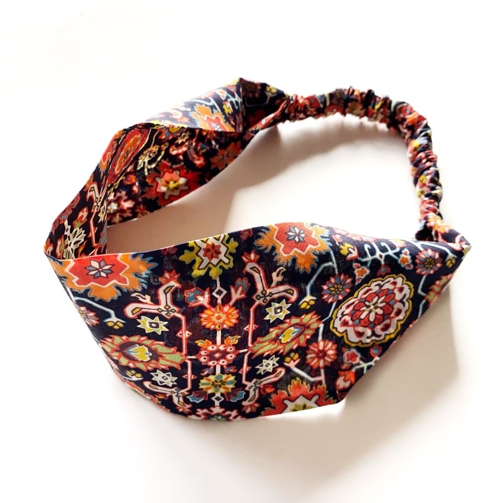 A Liberty London Tana Lawn cotton headband with a colorful floral and geometric pattern on a dark background, featuring an elastic band, photographed on a white background.