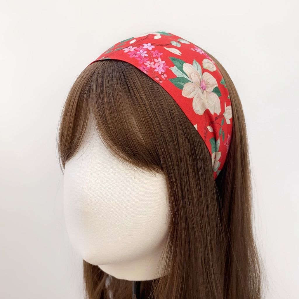 A orange red Liberty London Tana Lawn cotton headband with a floral pattern of pink and white flowers and green leaves, displayed on a mannequin head with long brown hair against a plain white background.