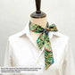 A tropical leaf print Liberty Tana Lawn cotton skinny scarf styled on a beige mannequin wearing a white collared shirt, against a light gray background. A text overlay at the bottom left explains that fabric patterns may vary.
