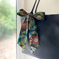 A Liberty Tana Lawn cotton skinny scarf featuring a green tropical leaf and deep red floral pattern, tied into a bow on a black handbag, shown hanging indoors next to a window.