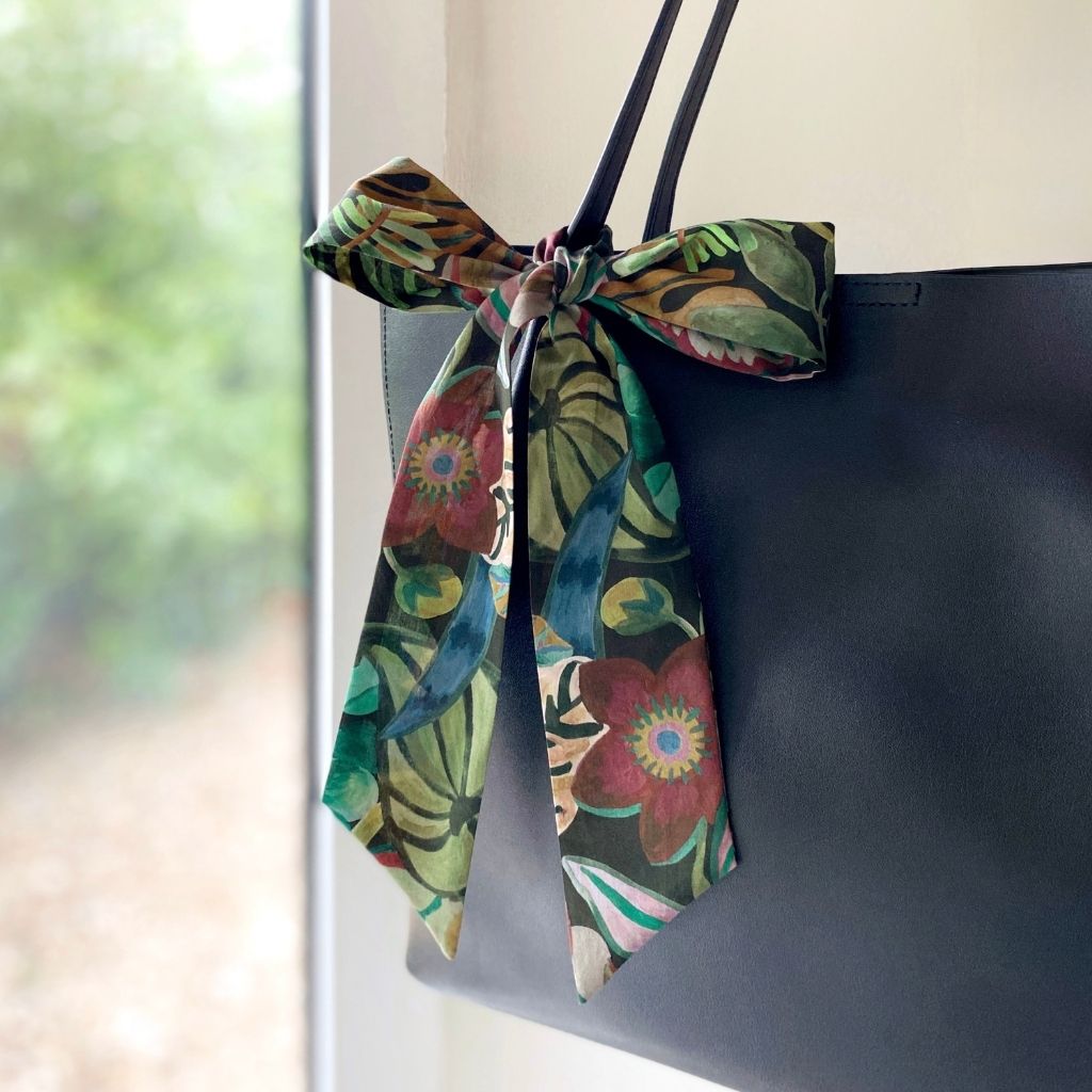 A Liberty Tana Lawn cotton skinny scarf featuring a green tropical leaf and deep red floral pattern, tied into a bow on a black handbag, shown hanging indoors next to a window.
