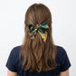 A woman viewed from the back wearing a navy blue T-shirt and a green Liberty Tana Lawn skinny hair scarf with a tropical leaf print, tied into a bow, set against a plain white background.