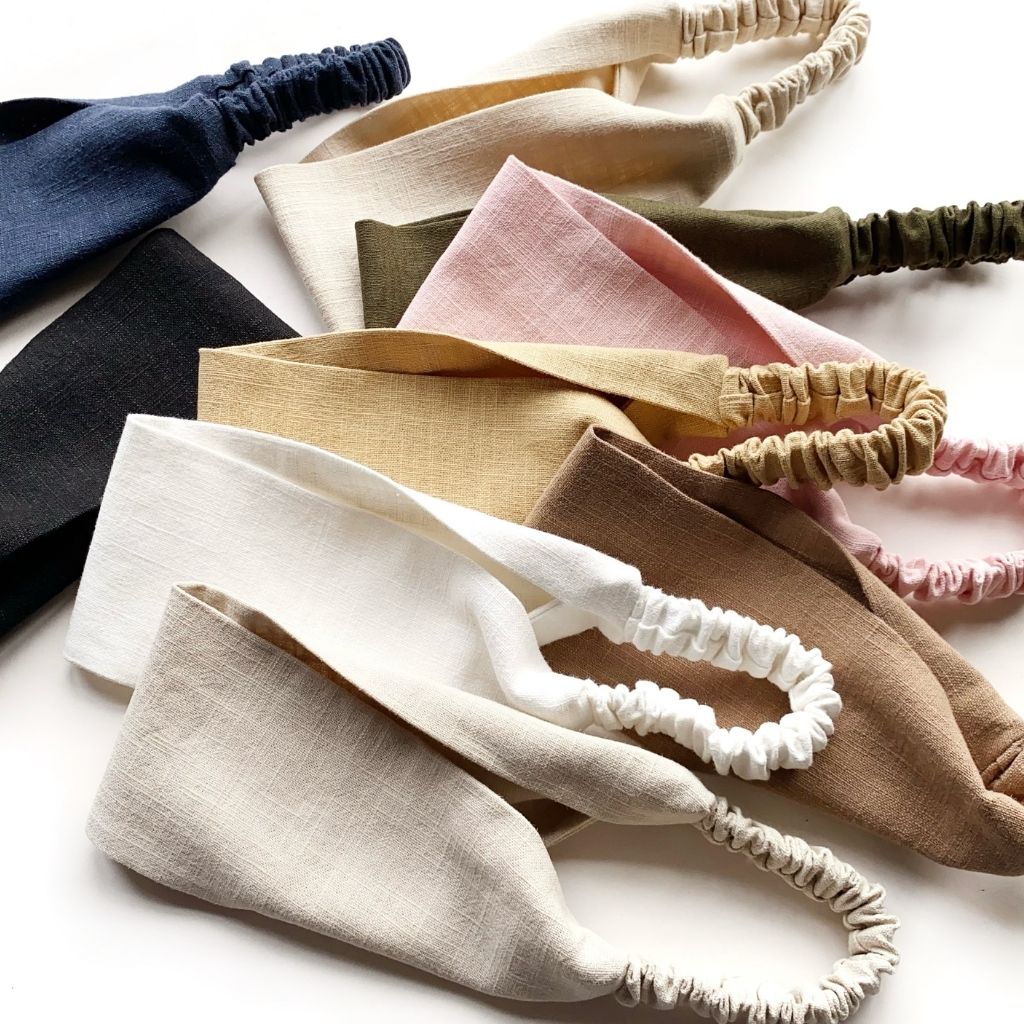 A set of linen headbands with gathered elastic back, shown in multiple soft colors including navy, black, cream, beige, brown, tan, pink, and olive green, arranged on a white background.