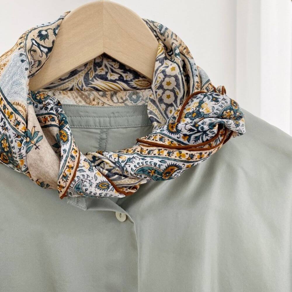 Yellow and teal paisley mulberry silk and cotton blend square neck scarf styled around the collar of a light green shirt on a hanger.