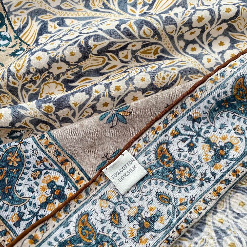 Close-up of yellow and teal paisley floral mulberry silk and cotton blend bandana square scarf showing detailed pattern and hand-rolled hem with fabric label.