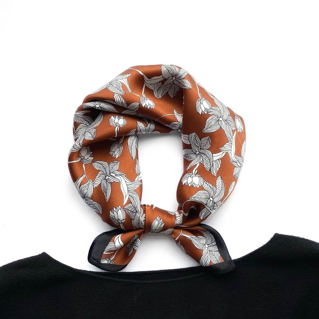 Brown line art floral pattern mulberry silk square scarf tied into a neck scarf, placed next to a black crewneck jumper on a white background.