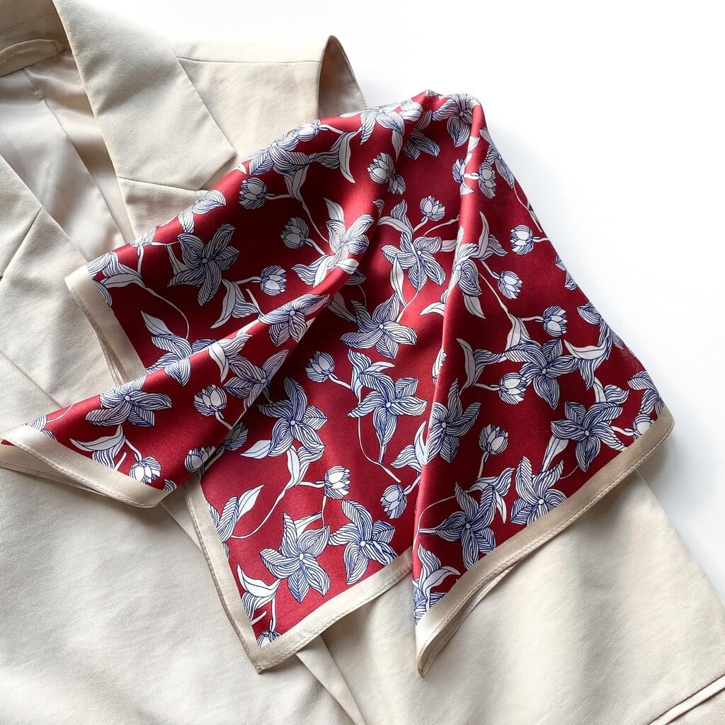 Folded mulberry silk square scarf with a navy and white line art floral pattern on a red background with a beige border, laid flat on a beige top.