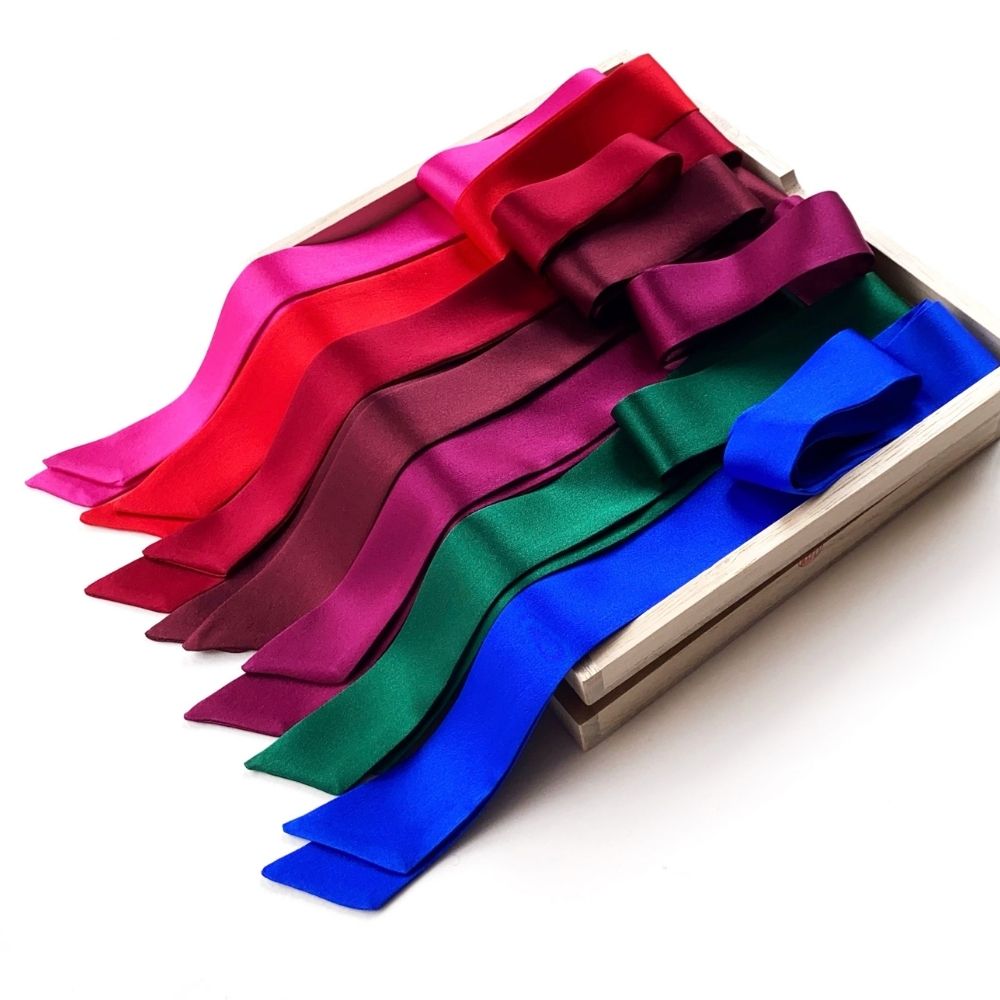 Seven mulberry silk satin scarves in a spectrum of colors including red, pink, purple, green, blue, and dark red, maroon, neatly arranged inside a light-colored wooden box on a white background.