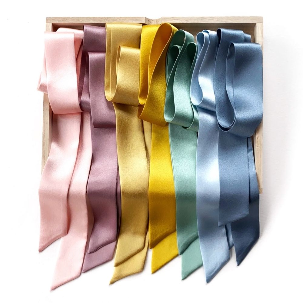Seven mulberry silk skinny scarves in pastel shades of pink, mauve, yellow gold, mint green, pastel blue and dusty blue, arranged in a light wood box on a white background.