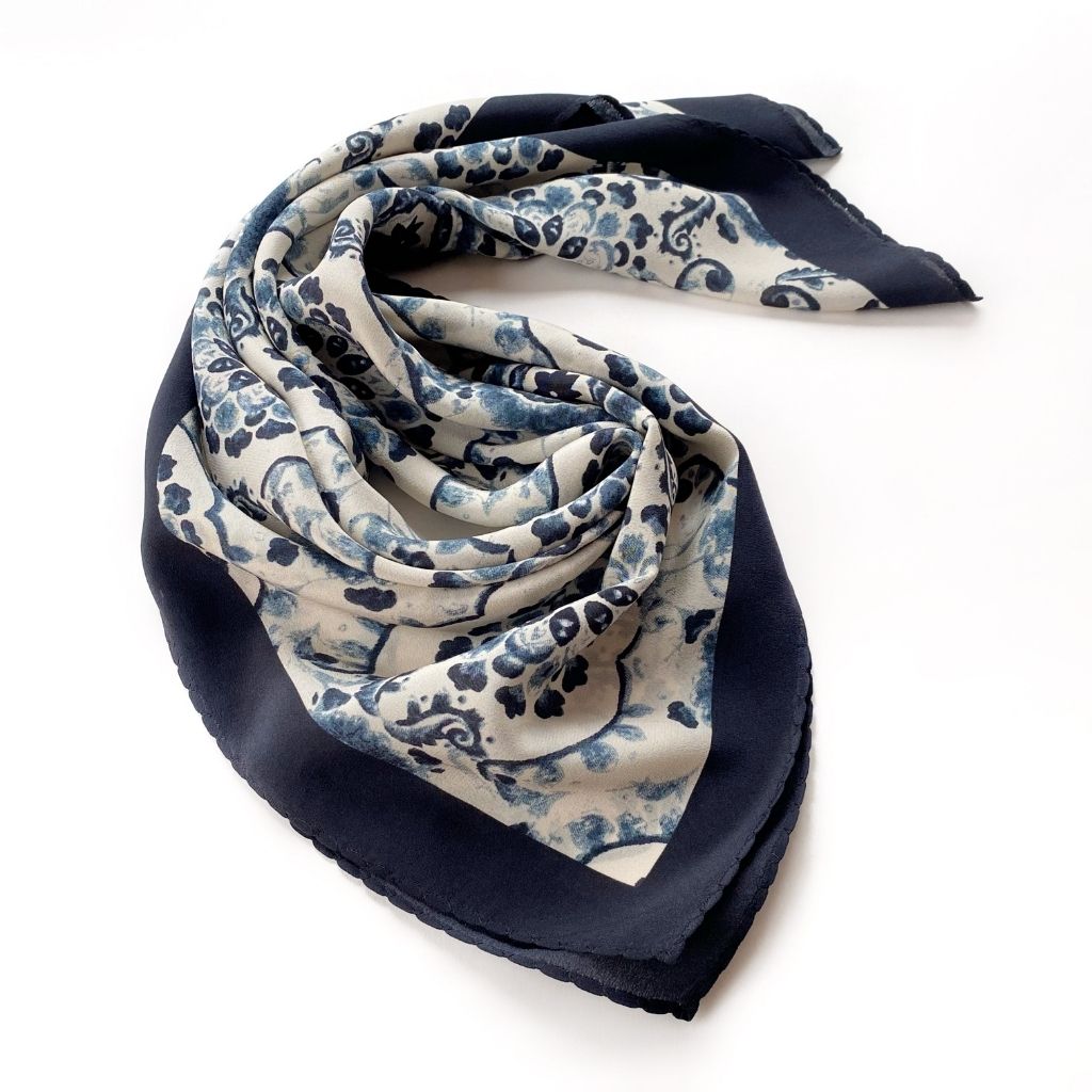 A rolled blue and white geometric floral print mulberry silk bandana scarf laid flat on a white background.