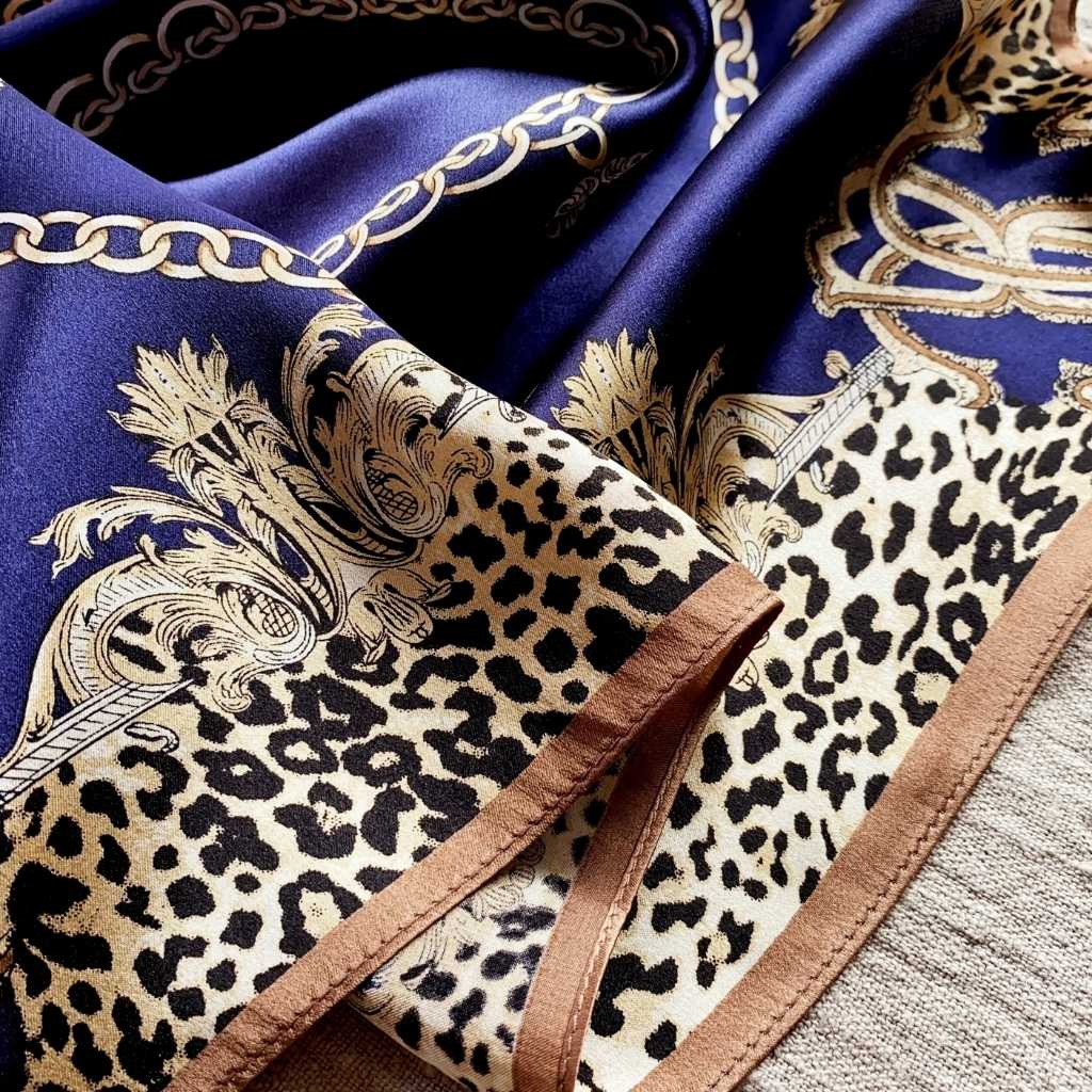 A close-up of a luxury mulberry silk scarf featuring a deep blue background with gold chain and baroque motifs, a beige leopard print section, and a contrast border.