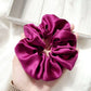 purple silk satin hair scrunchie held in a hand with a light wooden frame in the background