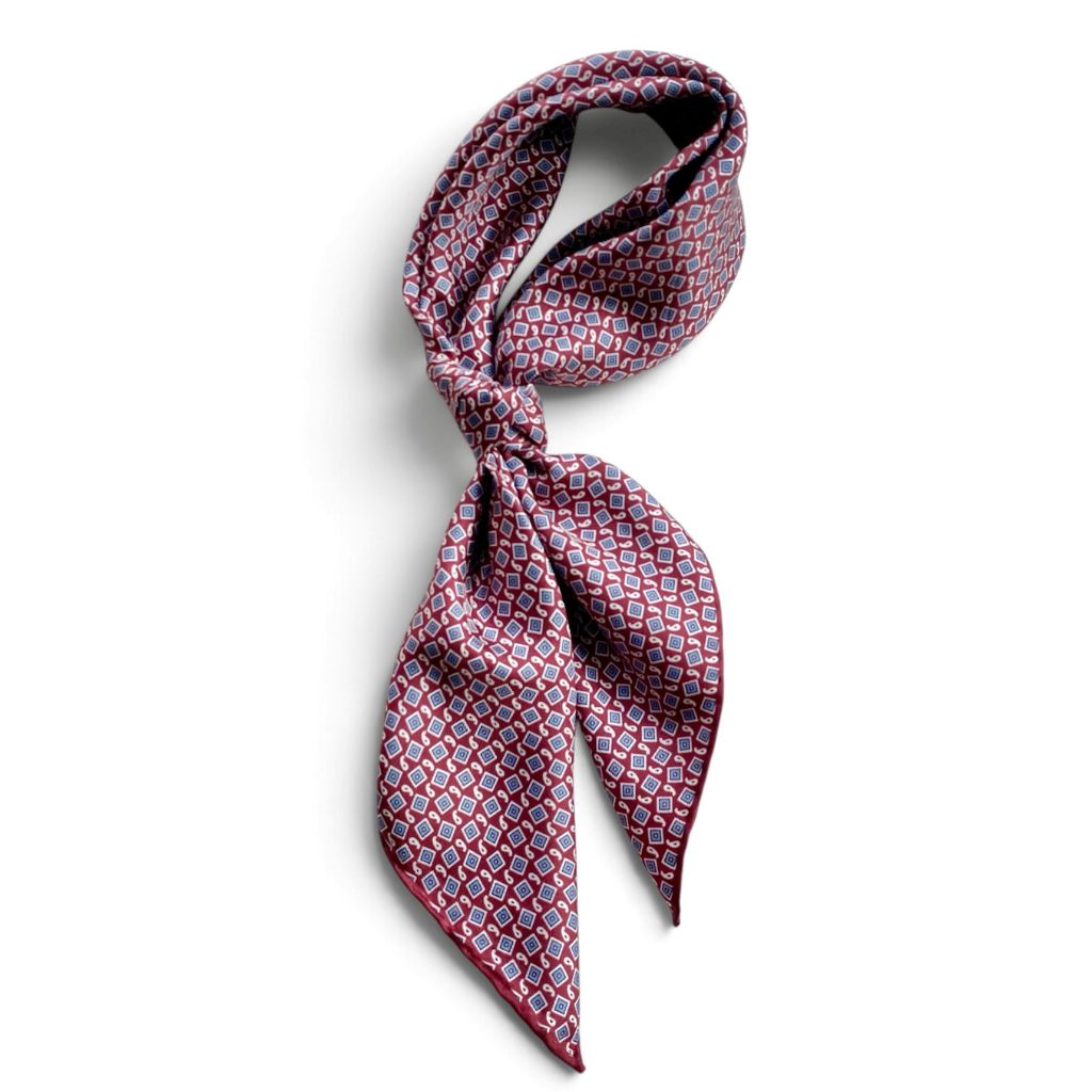 A patterned mulberry silk square scarf tied as a neckerchief, featuring a geometric and small paisley design in red, blue, and white on a white background