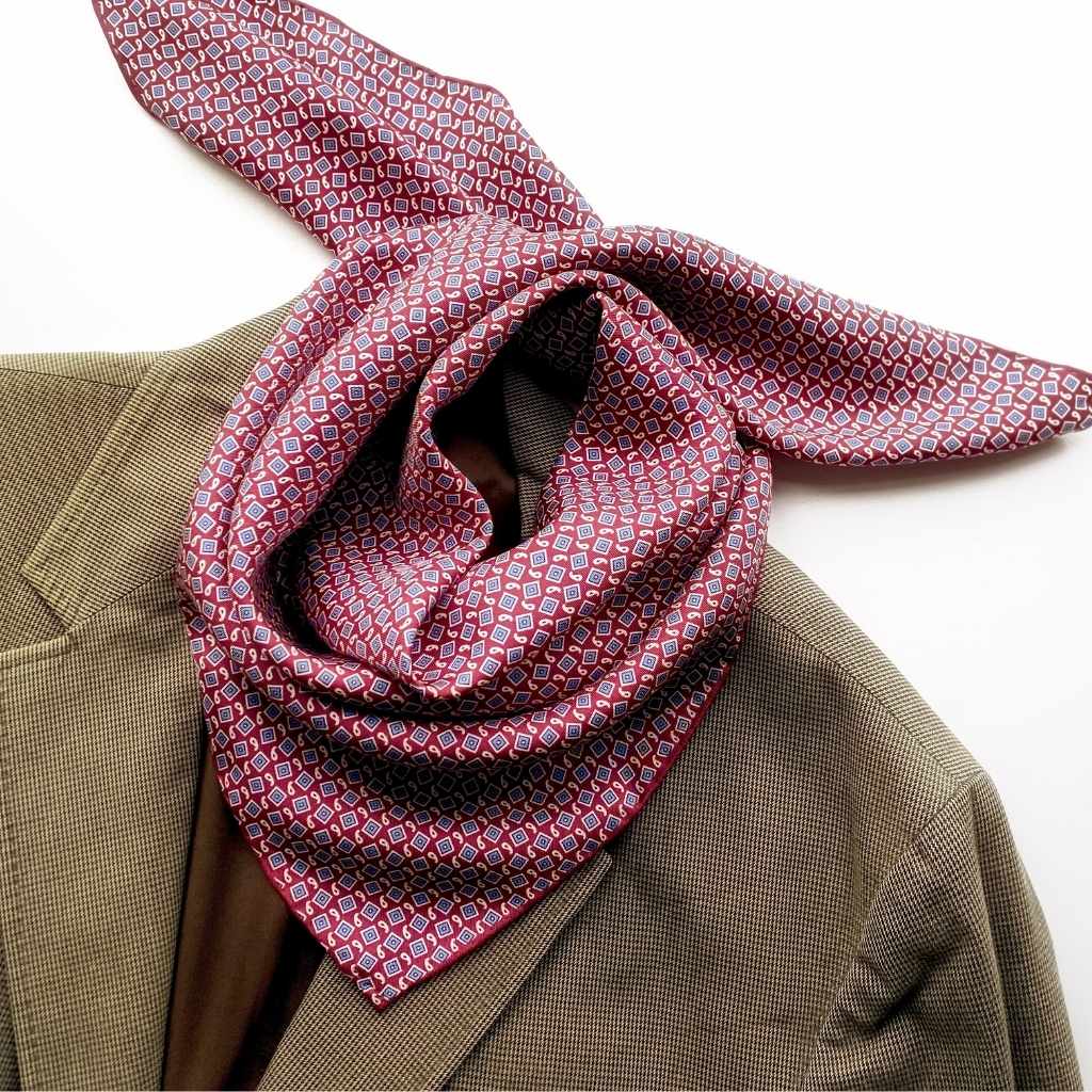 A patterned mulberry silk square scarf in red, blue, and white, tied around the collar of a brown tweed jacket against a plain white background
