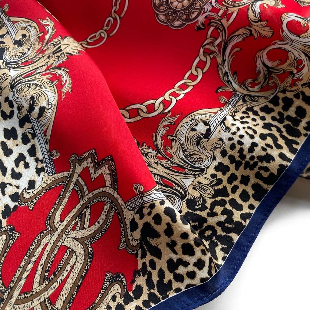 A close-up of a patterned mulberry silk square scarf featuring a bold red background with gold baroque scrollwork, leopard print sections, chain motifs, and a navy blue border.