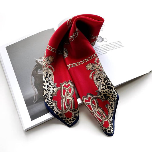 A red mulberry silk scarf with gold chain and baroque motifs, leopard print borders, and a navy blue frame, draped over an open book on a white background.