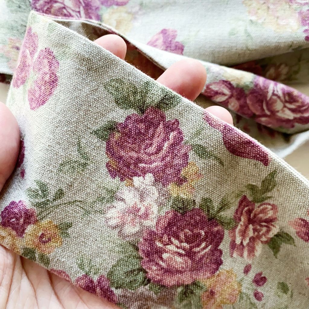 A close-up of a hand holding a rose floral print cotton linen headband with large mauve and pink roses, green leaves, and small yellow flowers on a light sage green background.