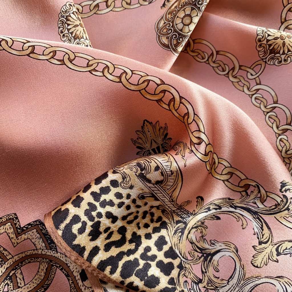 A close-up of a dusty rose mulberry silk scarf featuring a leopard print panel, ornate gold chain motifs, and baroque scrollwork.