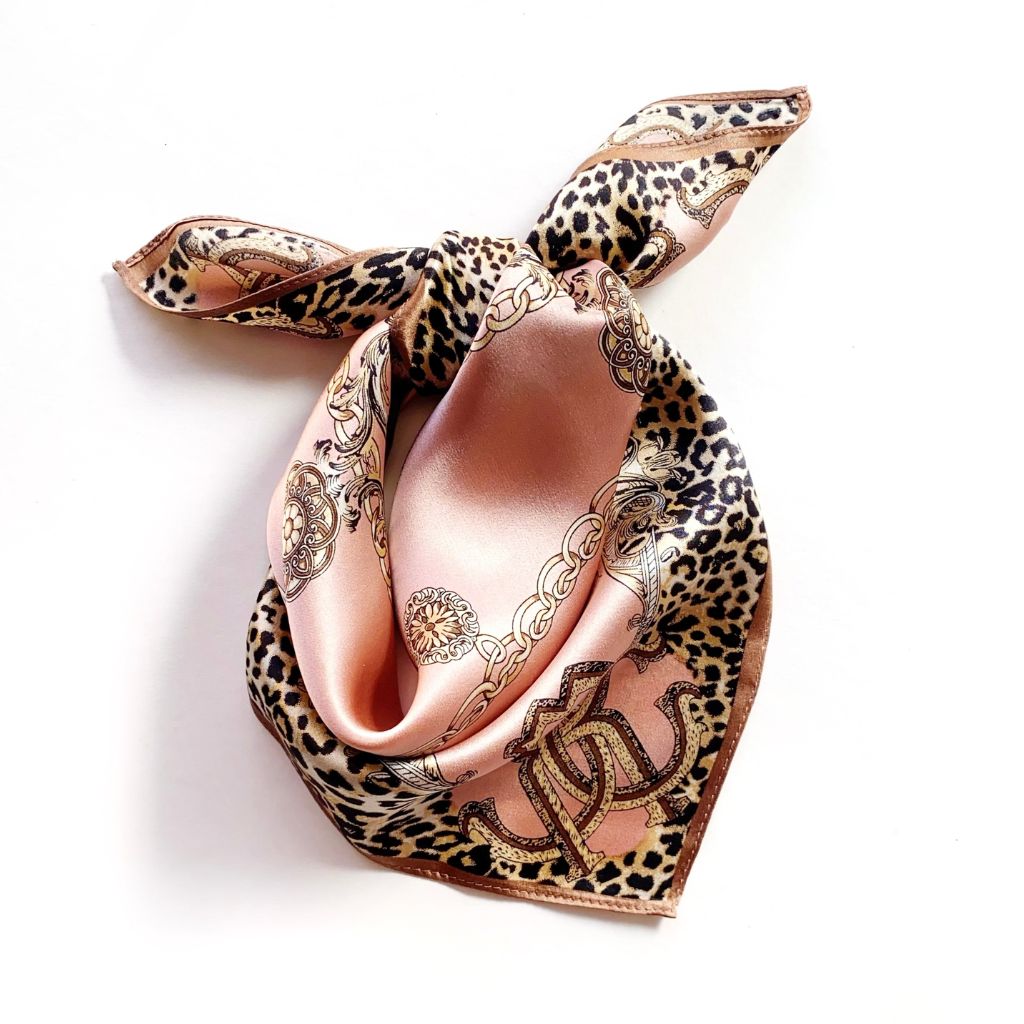 A square mulberry silk scarf with a leopard print border and a dusty pink center, featuring ornate gold chain and scroll patterns, tied in a knot on a white background.