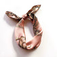A pink mulberry silk scarf with a gold and black baroque pattern, tied in a knot on a white background.