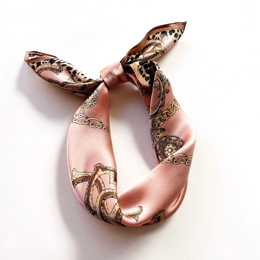 A pink mulberry silk scarf with a gold and black baroque pattern, tied in a knot on a white background.