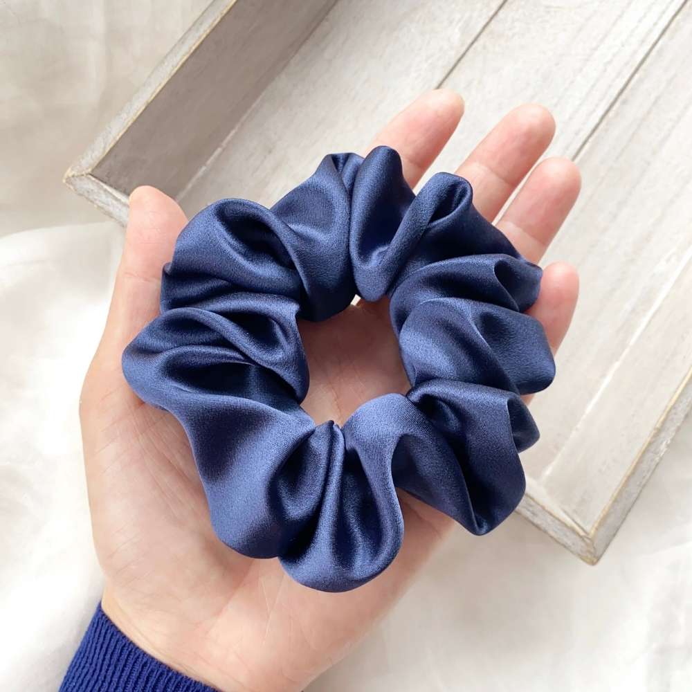 Hand holding a sapphire blue silk satin scrunchie on a light wooden surface