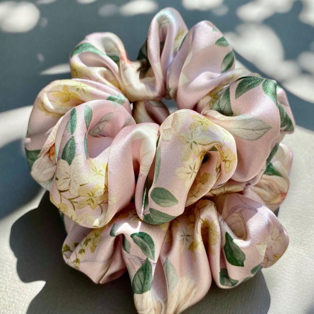 A liberty silk satin scrunchie with a light pink base and a floral print featuring green leaves and yellow flowers, resting on a neutral surface under dappled sunlight.