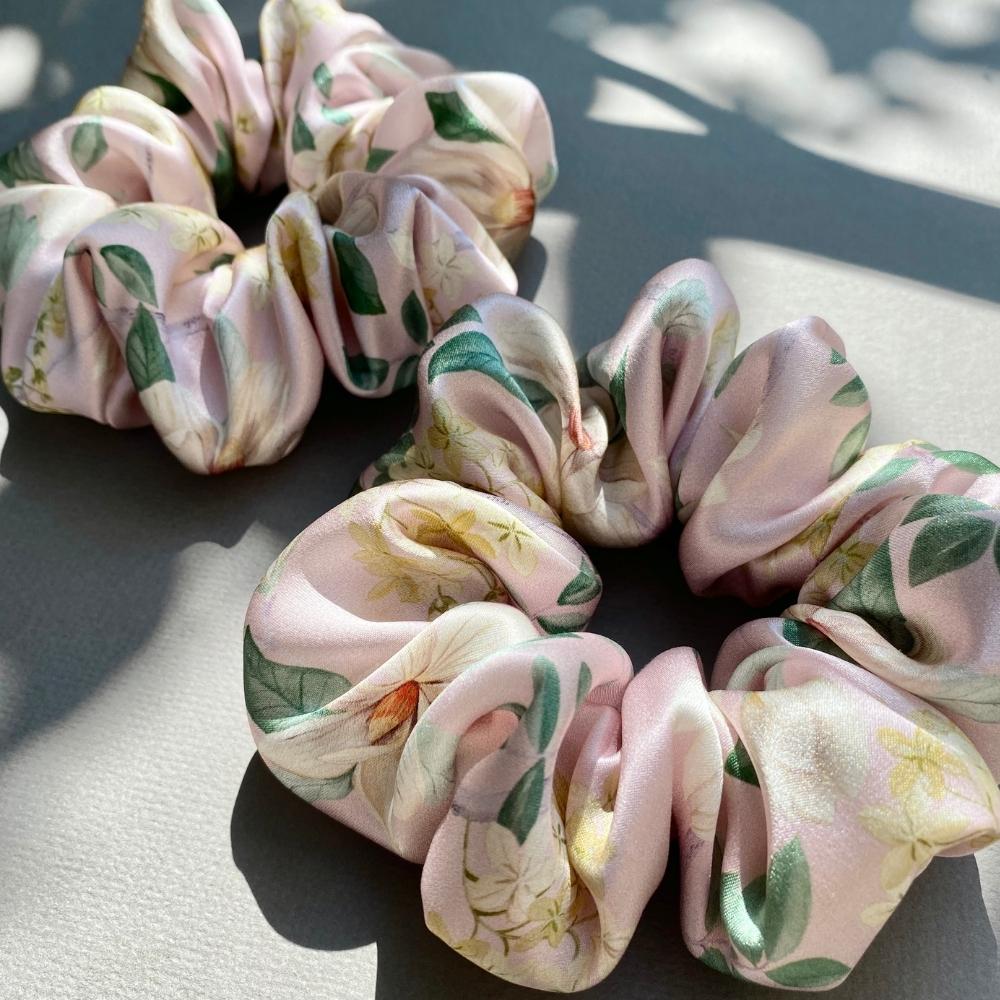 Two liberty silk satin hair scrunchies with a light pink floral print, featuring green leaves and yellow flowers, displayed on a neutral gray surface with soft shadows.