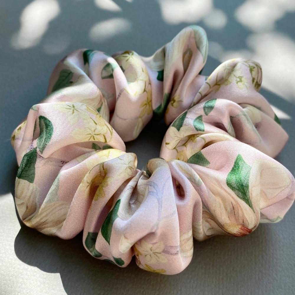 A liberty silk satin hair scrunchie with a soft pink base and a delicate floral print in cream, green, and yellow, photographed in natural dappled sunlight on a neutral gray surface.