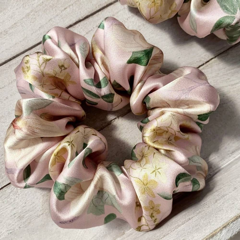 A liberty silk satin hair scrunchie with a light pink floral and green leaf pattern, displayed on a light-colored wooden surface.
