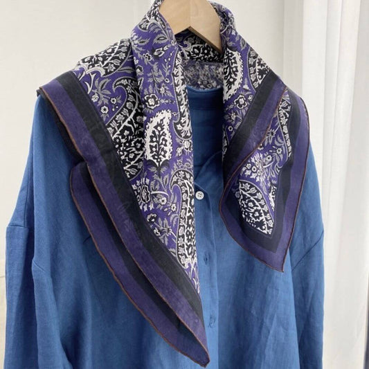 Purple paisley silk cotton blend square scarf draped over a blue shirt on a hanger, showing the hand-rolled hem and patterned border.