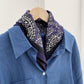 Purple paisley silk cotton blend scarf styled around the collar of a blue shirt on a hanger.