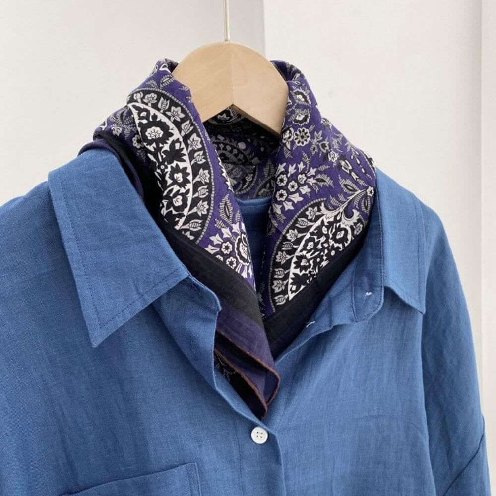 Purple paisley silk cotton blend scarf styled around the collar of a blue shirt on a hanger.